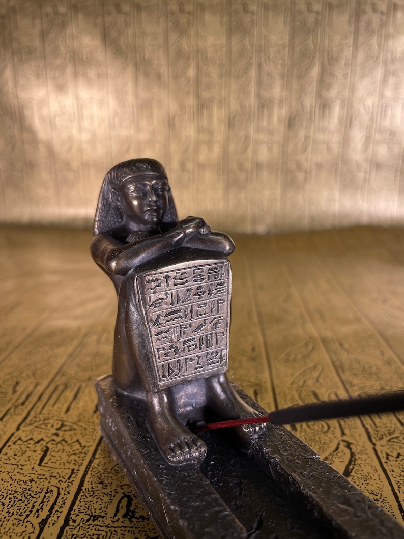 May include: Bronze incense burner shaped like an Egyptian pharaoh sitting with crossed legs. The burner has a rectangular incense holder with hieroglyphics carved into it. A stick of incense is burning in the holder.