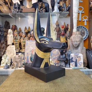 May include: Black Anubis statue with gold accents. The statue is a bust of the jackal-headed god Anubis, a prominent figure in ancient Egyptian mythology. The statue is displayed on a table with a gold patterned surface.