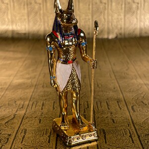 May include: A gold figurine of Anubis, the Egyptian god of the dead, with a jackal head, standing on a base. He is holding a staff in his right hand.
