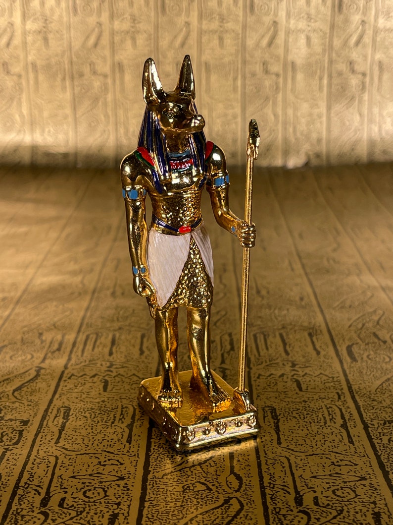 May include: A gold figurine of Anubis, the Egyptian god of the dead, with a jackal head, standing on a base. He is holding a staff in his right hand.
