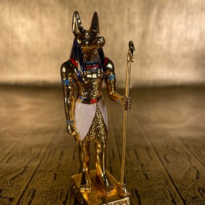 May include: A gold figurine of Anubis, the Egyptian god of the dead, with a jackal head, holding a staff. The figurine is standing on a gold base.