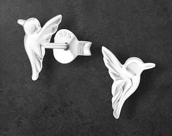 Könnte beinhalten: Ein Paar Ohrstecker aus Sterlingsilber mit Kolibri-Motiv. Die Ohrringe sind in Form eines Kolibris mit detaillierten Flügeln und Körpern gestaltet. Der Stift eines Ohrrings ist sichtbar, mit "925" gestempelt. Dunkler Hintergrund.