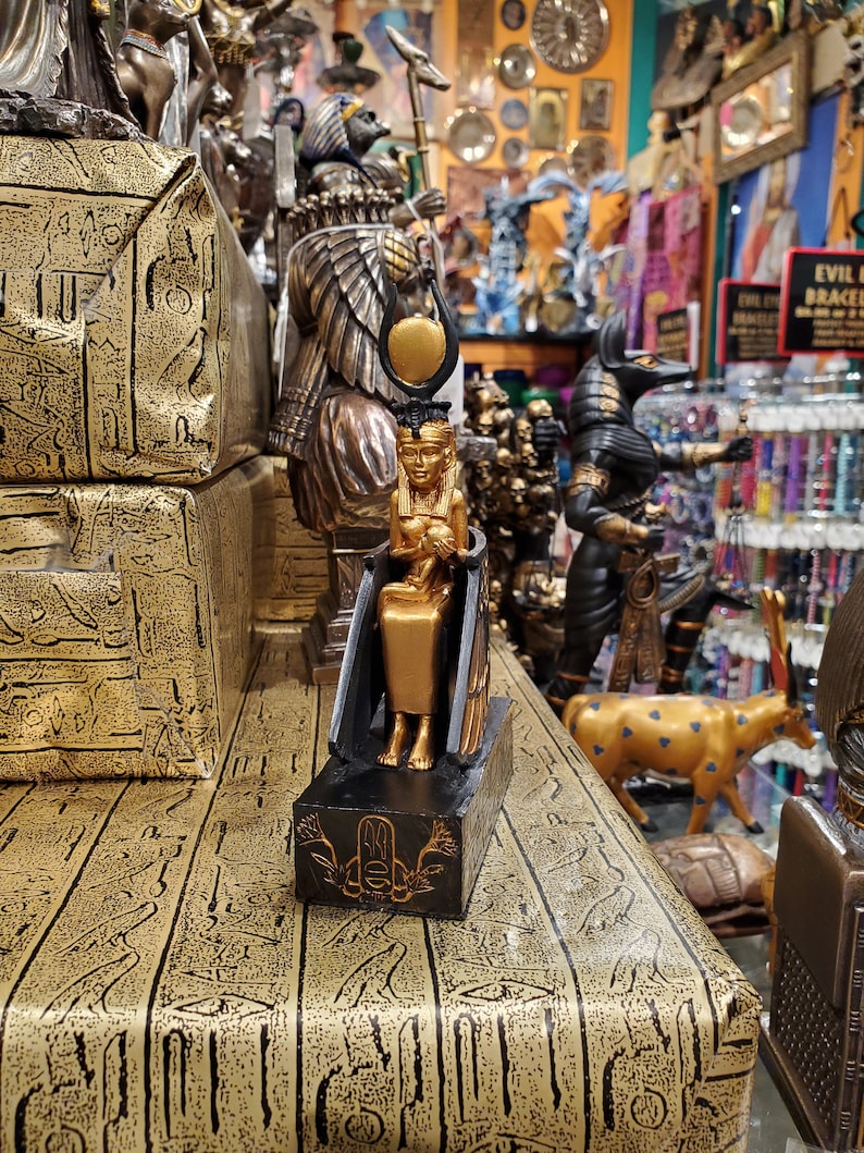 May include: Gold and black figurine of an Egyptian goddess sitting on a throne. The goddess is wearing a crown and holding a small figure in her lap. The figurine is on a black base with a gold design.