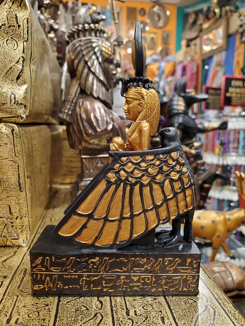 May include: Gold and black figurine of the Egyptian goddess Isis, seated on a throne with a winged figure, with hieroglyphics on the base.