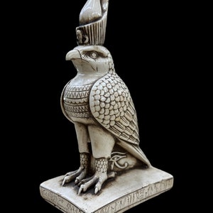 K&ouml;nnte beinhalten: Eine detaillierte, cremefarbene Statue eines &auml;gyptischen Falkengottes, m&ouml;glicherweise Horus, mit hoher Krone und aufwendigen Federdetails. Die Statue steht auf einem rechteckigen Sockel mit Hieroglyphen, vor schwarzem Hintergrund.