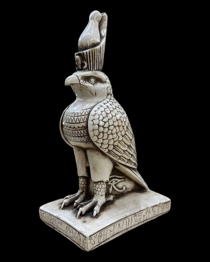 K&ouml;nnte beinhalten: Eine detaillierte, cremefarbene Statue eines &auml;gyptischen Falkengottes, m&ouml;glicherweise Horus, mit hoher Krone und aufwendigen Federdetails. Die Statue steht auf einem rechteckigen Sockel mit Hieroglyphen, vor schwarzem Hintergrund.
