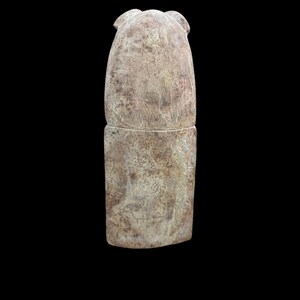 May include: A carved stone sculpture of a seated figure with a rounded head and a rectangular base. The sculpture is made of a light brown stone with a rough texture.