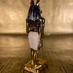 May include: A gold-colored figurine of the Egyptian god Anubis, with blue and white accents. The figurine is standing on a gold-colored base.