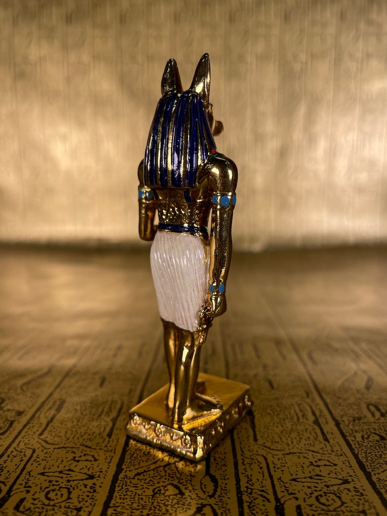 May include: A gold-colored figurine of the Egyptian god Anubis, with blue and white accents. The figurine is standing on a gold-colored base.