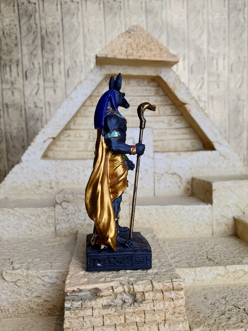 May include: Black and gold figurine of the Egyptian god Anubis standing on a black base with hieroglyphics. Anubis is holding a golden staff with a snake head.