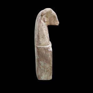 May include: A carved stone sculpture of a ram's head with a long neck. The sculpture is made of a light brown stone with a rough texture.