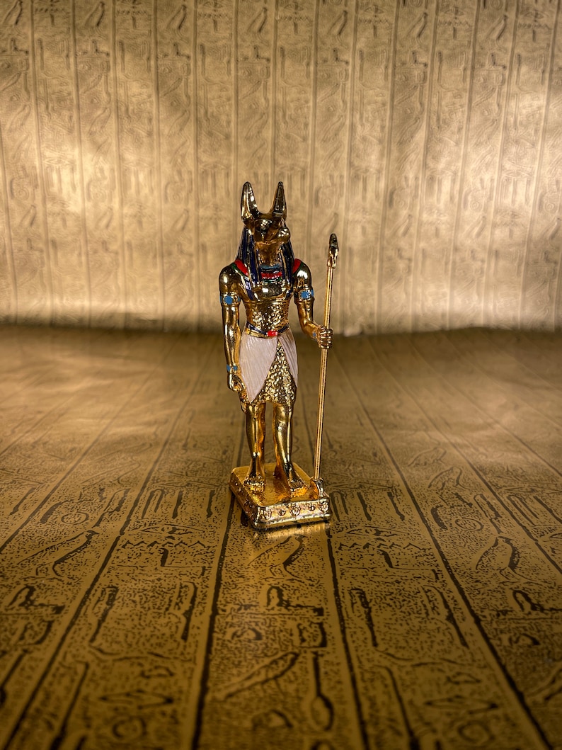 May include: A gold-colored figurine of the Egyptian god Anubis, standing on a base with a staff in his right hand. The figurine is detailed with blue and red accents.