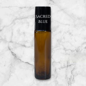 Op de afbeelding: Een bruine glazen rollerfles met een zwarte dop en een wit etiket met de tekst "SACRED BLUE".