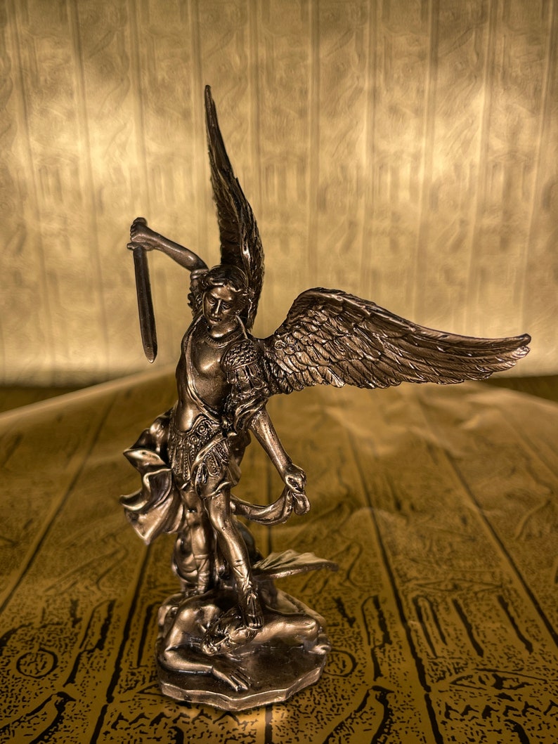 May include: A bronze statue of an angel with wings outstretched, holding a sword in one hand and standing over a fallen figure. The statue is on a brown patterned surface.