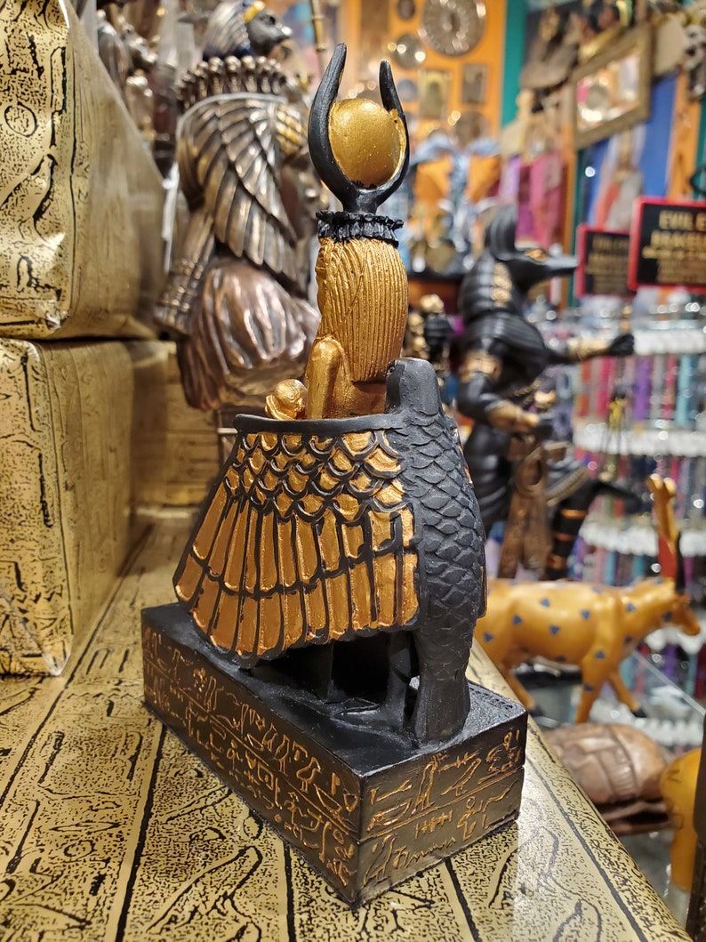 May include: A gold and black figurine of the Egyptian goddess Bastet, depicted as a cat with a human head and a sun disk on her head. The figurine is standing on a base with hieroglyphics.