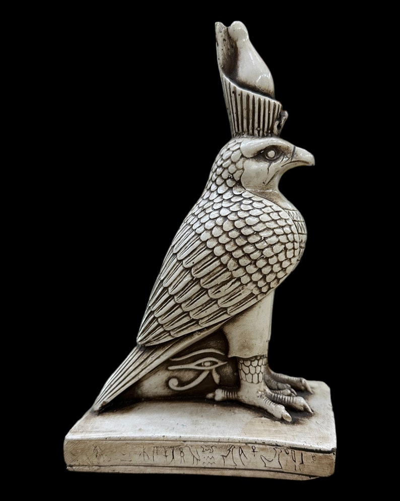 K&ouml;nnte beinhalten: Eine detaillierte, cremefarbene Statue einer falkenk&ouml;pfigen Gottheit, m&ouml;glicherweise Horus, mit aufwendigen Feder- und Schuppendetails. Die Statue steht auf einem rechteckigen Sockel mit Hieroglyphen und dem Horusauge.