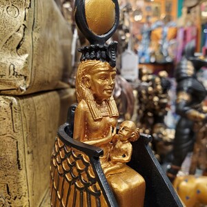 May include: Gold and black statue of an Egyptian goddess holding a child. The goddess is wearing a crown with a sun disc and has wings. The statue is on a black base.