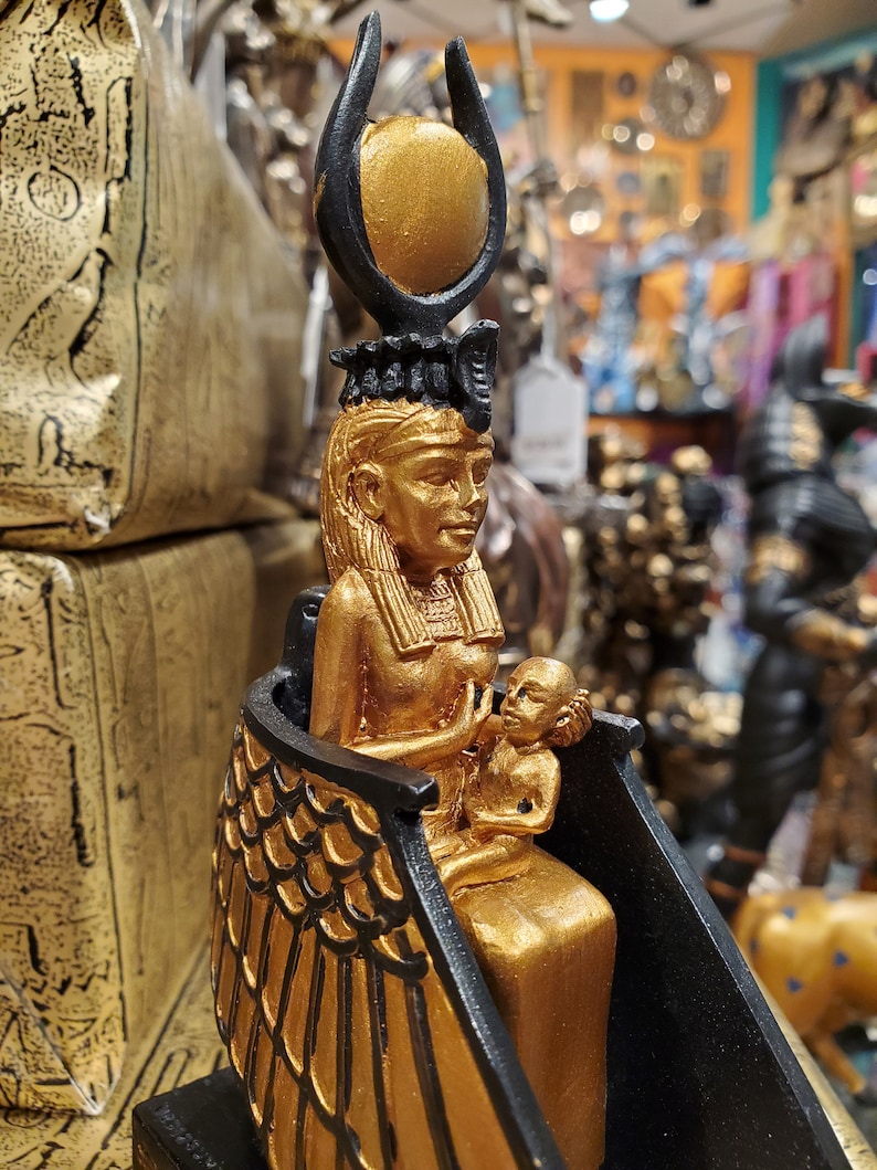 May include: Gold and black statue of an Egyptian goddess holding a child. The goddess is wearing a crown with a sun disc and has wings. The statue is on a black base.