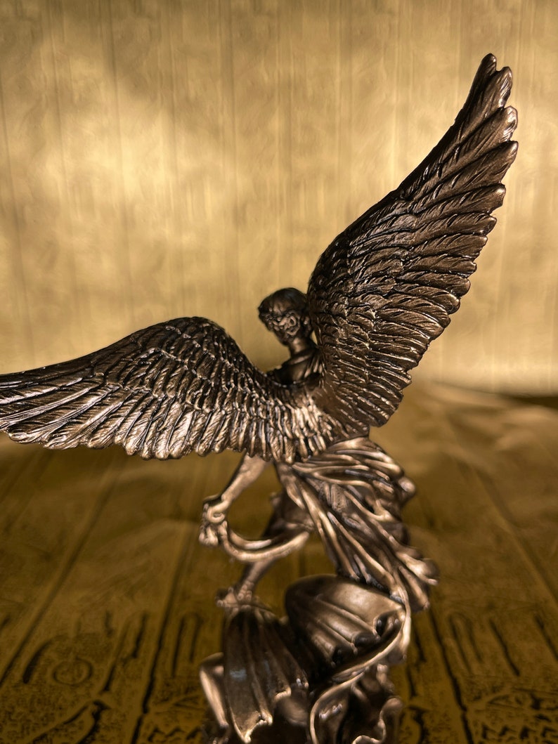 May include: A bronze statue of an angel with wings spread wide. The angel is standing on a base and is holding a piece of fabric in its hands.