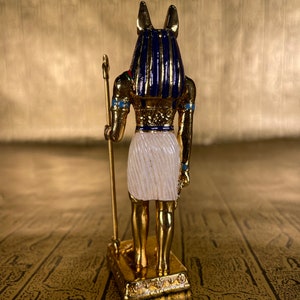 May include: A gold figurine of the Egyptian god Anubis, with a jackal head, holding a staff. The figurine is standing on a gold base and is painted with blue, white, and red accents.