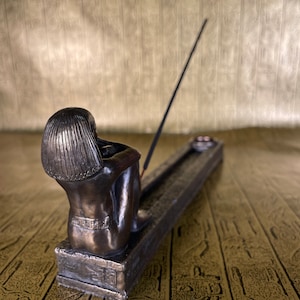 May include: Bronze incense burner featuring a seated figure with a detailed design. The burner has a long, thin slot for incense sticks and a small, round dish for ashes.