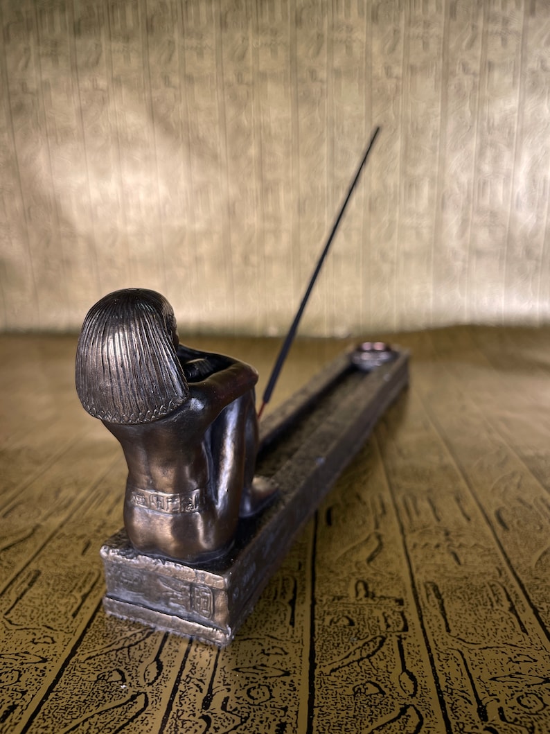 May include: Bronze incense burner featuring a seated figure with a detailed design. The burner has a long, thin slot for incense sticks and a small, round dish for ashes.