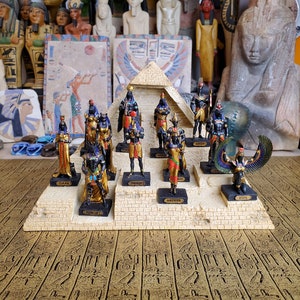 May include: A pyramid-shaped display stand with 10 small gold and black figurines of Egyptian gods and goddesses. Each figurine is on a small platform with the name of the god or goddess written below.
