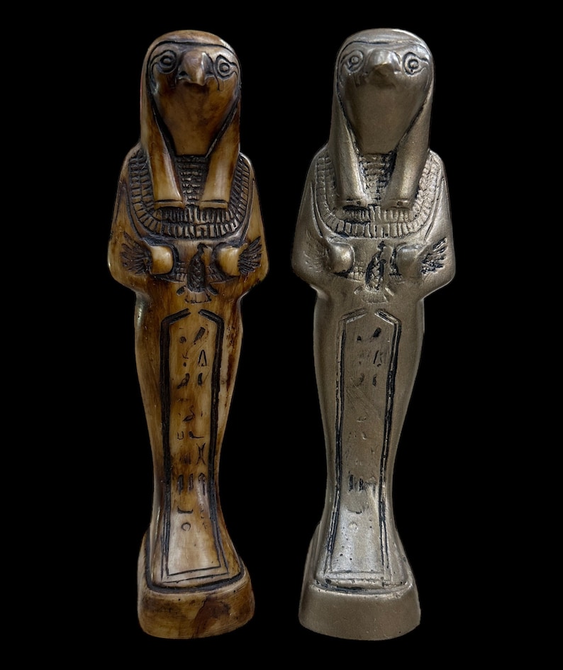May include: Two small statues of the Egyptian god Horus, one in a brown wood finish and the other in a silver finish. Both statues are standing with their arms crossed and have a hawk head.