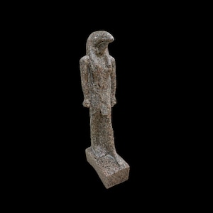 Puede incluir: Una estatua de piedra gris de un dios egipcio con cuerpo humano y cabeza de pájaro. La estatua está de pie sobre una base rectangular.