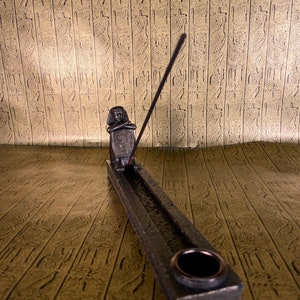 May include: A black metal incense burner with a small Egyptian figure holding a scroll. The burner has a groove for incense sticks and a round hole for ash.