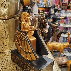 May include: Gold and black statue of the Egyptian goddess Isis holding a baby Horus. The statue is sitting on a black base with hieroglyphics.