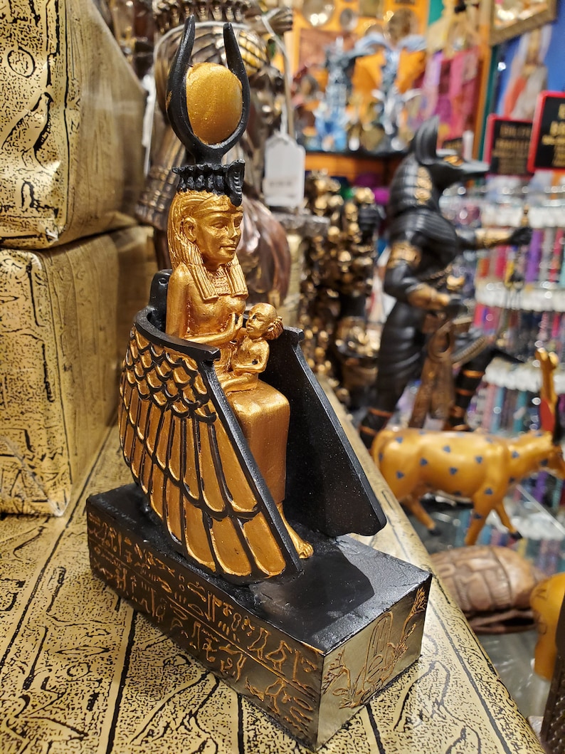 May include: Gold and black statue of the Egyptian goddess Isis holding a baby Horus. The statue is sitting on a black base with hieroglyphics.