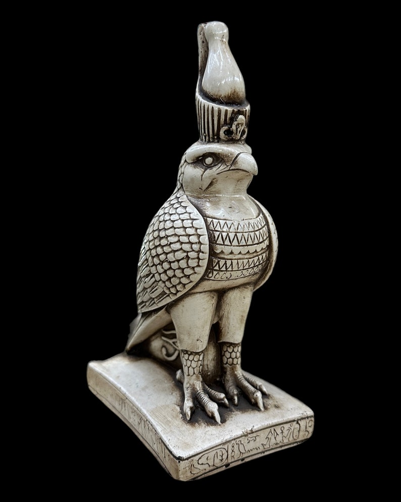 K&ouml;nnte beinhalten: Eine detaillierte, cremefarbene Statue einer falkenk&ouml;pfigen Gottheit, m&ouml;glicherweise Horus, mit aufwendigen Schnitzereien. Die Figur steht auf einem rechteckigen Sockel mit Hieroglyphen, vor schwarzem Hintergrund. Die Statue hat eine hohe Krone und detaillierte Federmuster.