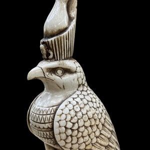 K&ouml;nnte beinhalten: Elfenbeinfarbene Statue einer falkenk&ouml;pfigen Gottheit, m&ouml;glicherweise Horus, mit einer hohen Kopfbedeckung. Die Skulptur weist detaillierte Schnitzereien von Federn und komplizierten Mustern auf der Brust auf, vor schwarzem Hintergrund.