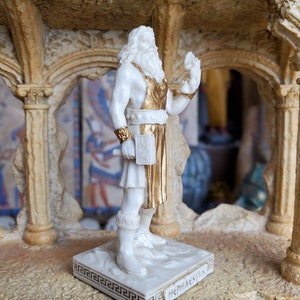 May include: White figurine of the Greek god Hephaestus, holding a hammer and wearing a gold-trimmed apron. The figurine is standing on a white base with a Greek key border and the inscription "Hephaestus".