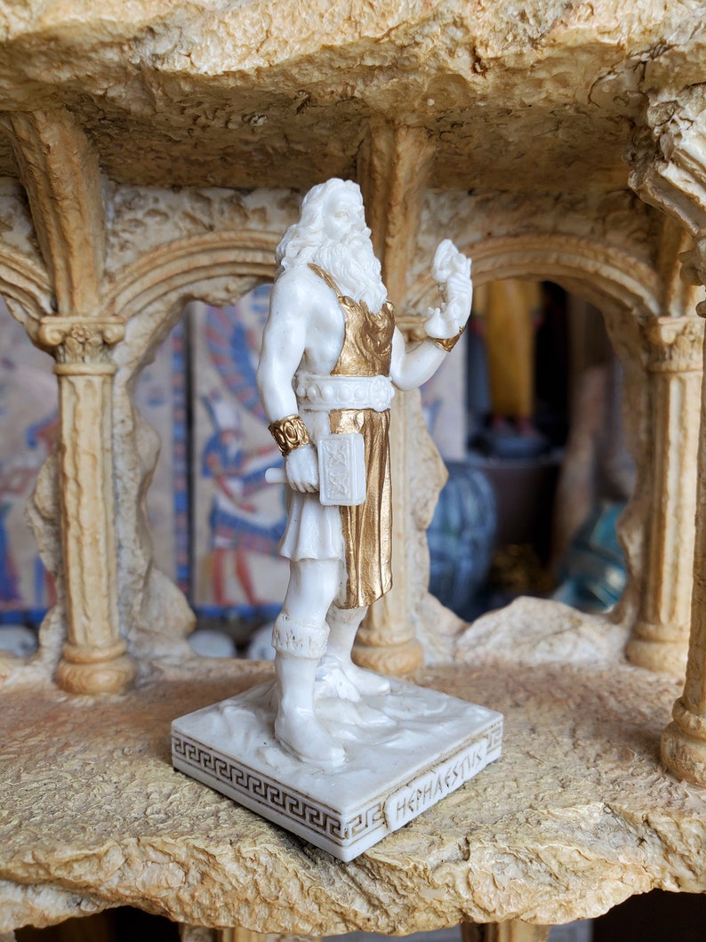 May include: White figurine of the Greek god Hephaestus, holding a hammer and wearing a gold-trimmed apron. The figurine is standing on a white base with a Greek key border and the inscription "Hephaestus".