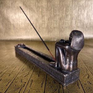 May include: A bronze incense burner shaped like a seated figure with a long incense stick burning. The burner is on a textured surface.