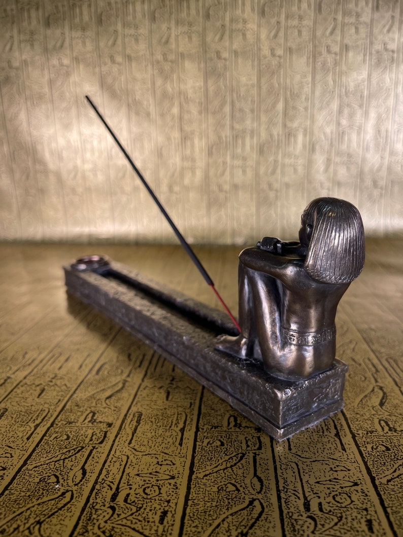 May include: A bronze incense burner shaped like a seated figure with a long incense stick burning. The burner is on a textured surface.