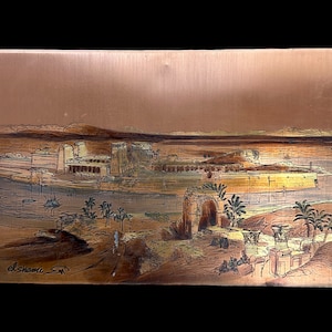 May include: A copper plaque with an image of an ancient Egyptian temple complex. The temple is depicted in a desert setting with palm trees and a river in the foreground. The plaque is signed "el shami" in the lower left corner.