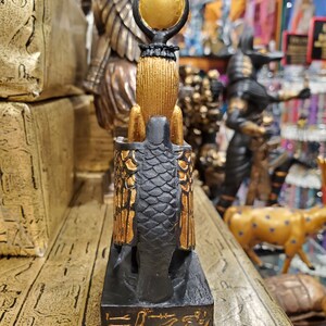 May include: A black and gold figurine of the Egyptian goddess Bastet, depicted as a cat with a human head and wings. The figurine is standing on a black base with hieroglyphs inscribed in gold.