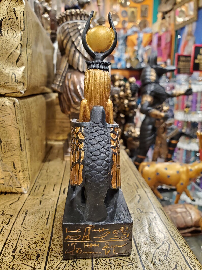 May include: A black and gold figurine of the Egyptian goddess Bastet, depicted as a cat with a human head and wings. The figurine is standing on a black base with hieroglyphs inscribed in gold.