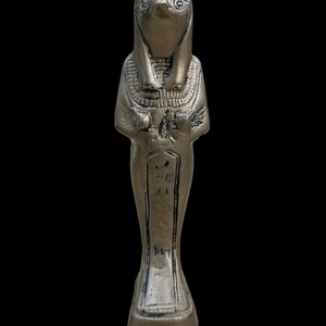 May include: Silver-colored figurine of an Egyptian god with a bird head, wearing a traditional headdress and holding a staff. The figurine is standing on a rectangular base.