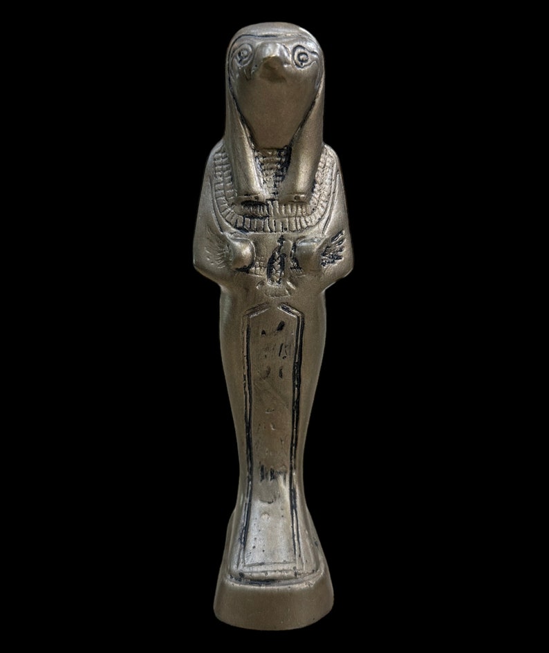 May include: Silver-colored figurine of an Egyptian god with a bird head, wearing a traditional headdress and holding a staff. The figurine is standing on a rectangular base.