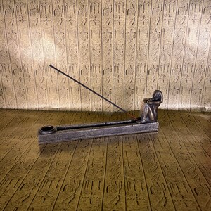 May include: A bronze incense burner shaped like a seated figure with a stick of incense burning. The burner is on a textured surface.