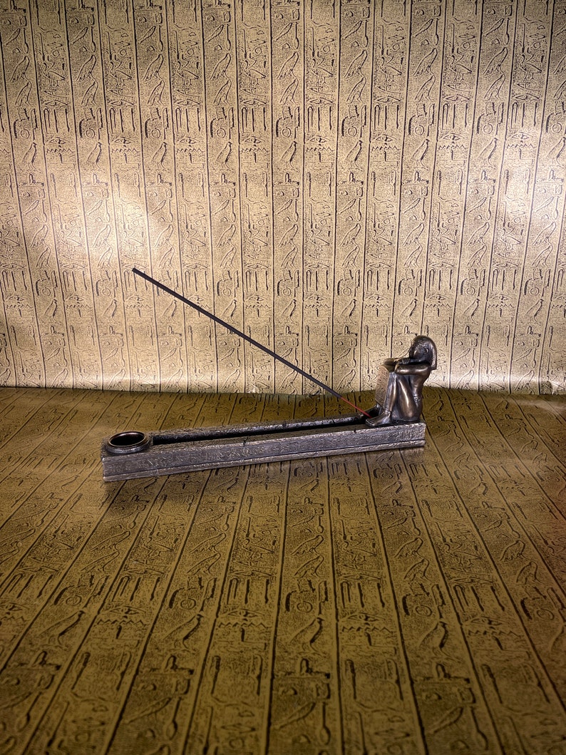 May include: A bronze incense burner shaped like a seated figure with a stick of incense burning. The burner is on a textured surface.