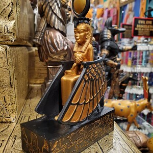 May include: Gold and black figurine of the Egyptian goddess Isis, sitting on a throne with wings, wearing a crown with a sun disc. The base of the figurine is decorated with hieroglyphs.