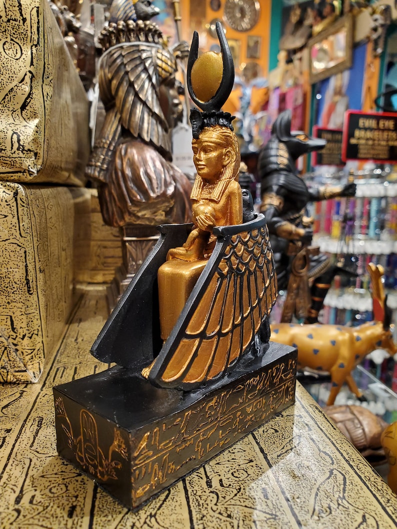 May include: Gold and black figurine of the Egyptian goddess Isis, sitting on a throne with wings, wearing a crown with a sun disc. The base of the figurine is decorated with hieroglyphs.