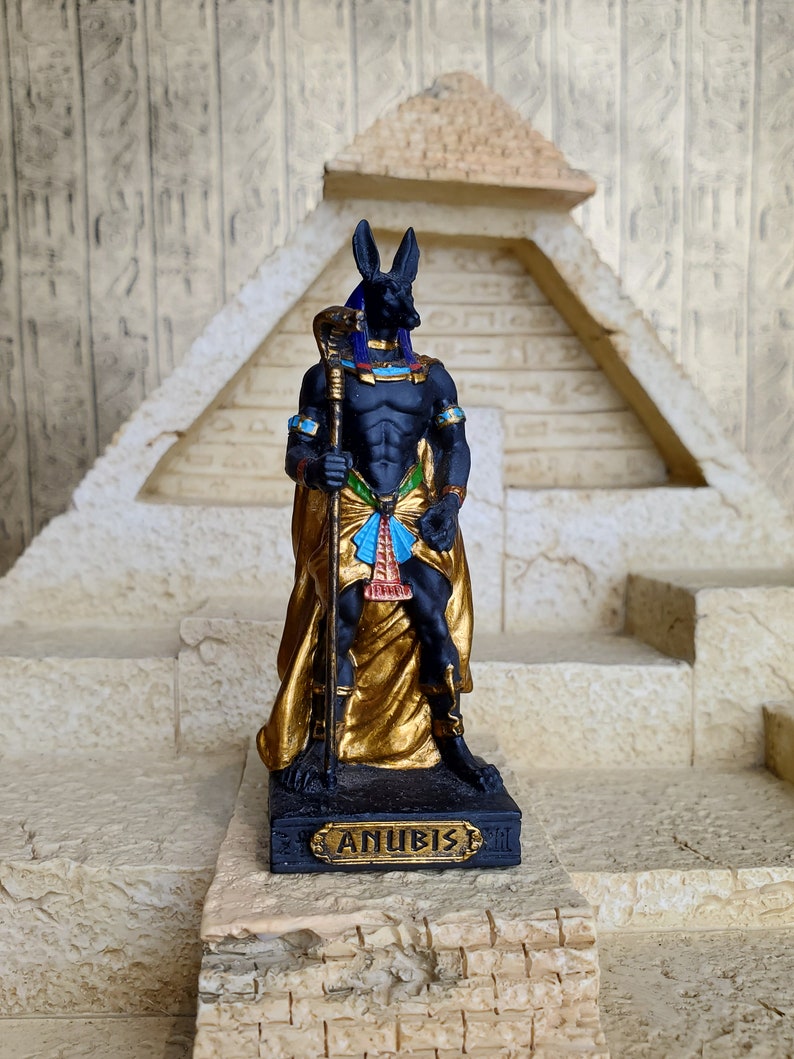 May include: Black Anubis statue with a gold and blue robe, holding a staff. The statue is standing on a base with the word "Anubis" inscribed on it.