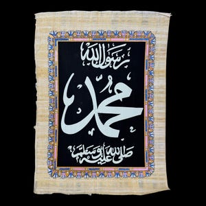 May include: A framed Islamic calligraphy artwork on papyrus paper with a black background and white Arabic script. The script reads "Allah" and "Muhammad" in Arabic. The frame is decorated with a colourful floral pattern.