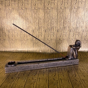 May include: A bronze incense burner with an Egyptian figure sitting on the side. The burner has a rectangular shape with a groove for incense sticks and a small round hole for ash. The figure is holding a hand to its chin and is looking down.